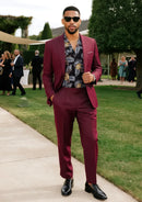 Man in a burgundy suit with a floral shirt standing outdoors on a grassy area.
